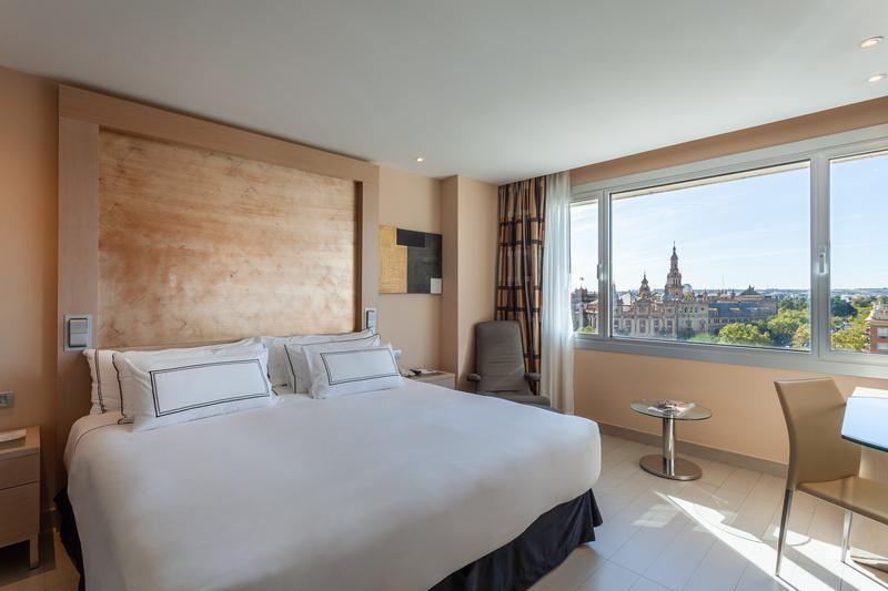 Modern hotel room with a large bed and view of the city through a wide window.