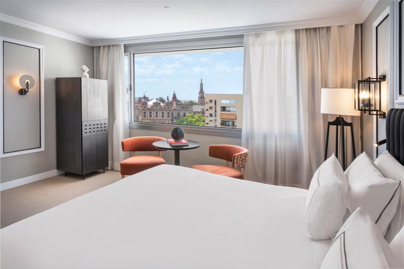 Modern hotel room with a large bed and view of the city through a wide window.
